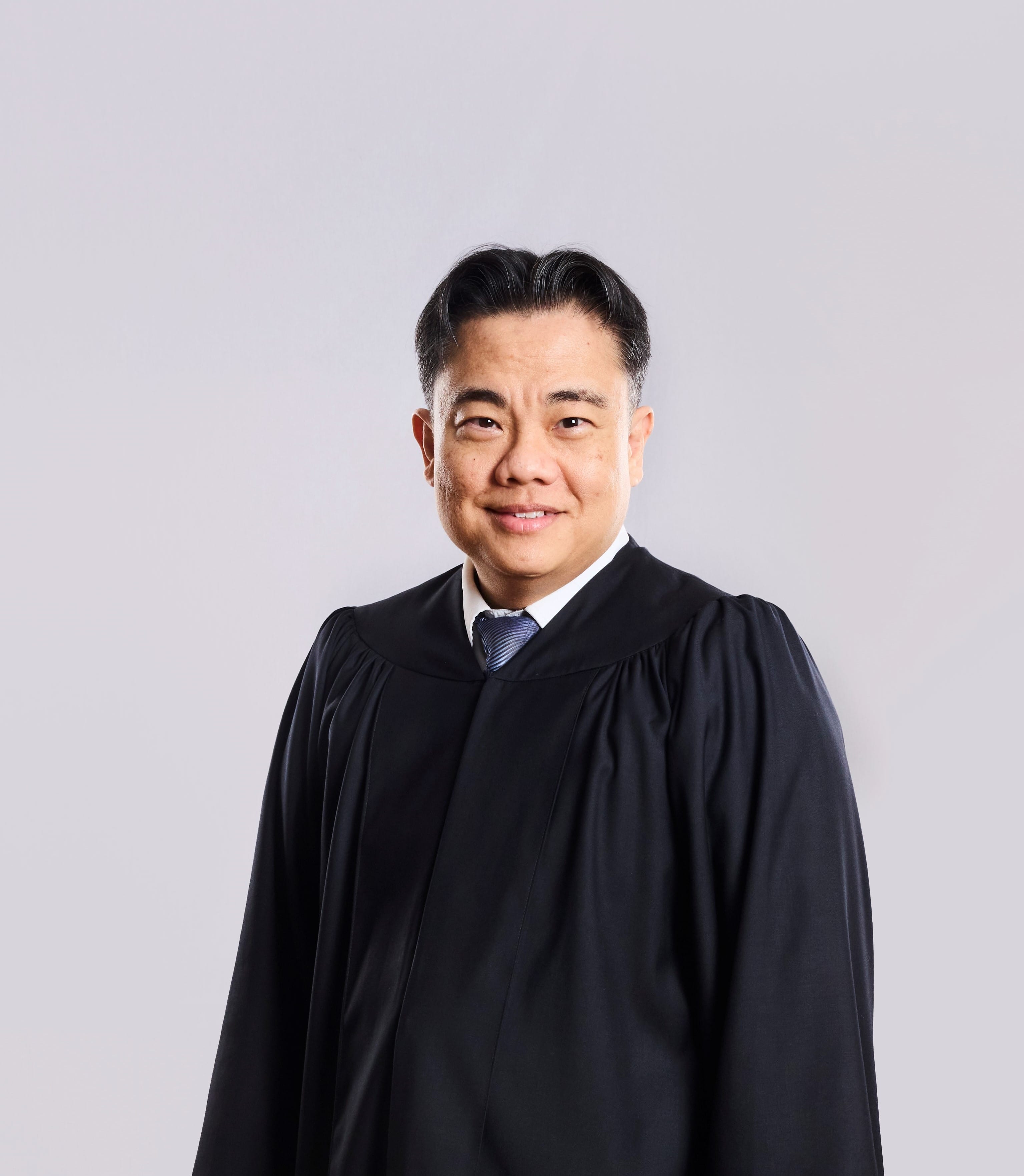 Person in dark robe, white shirt, and striped tie against a light gray background.